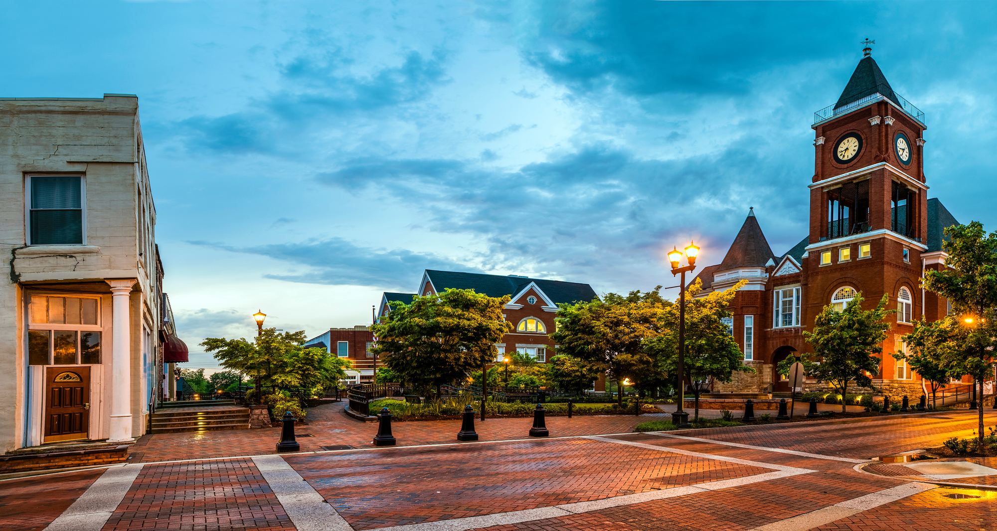 A small town square at dusk with a brick clock tower and lit street lamps