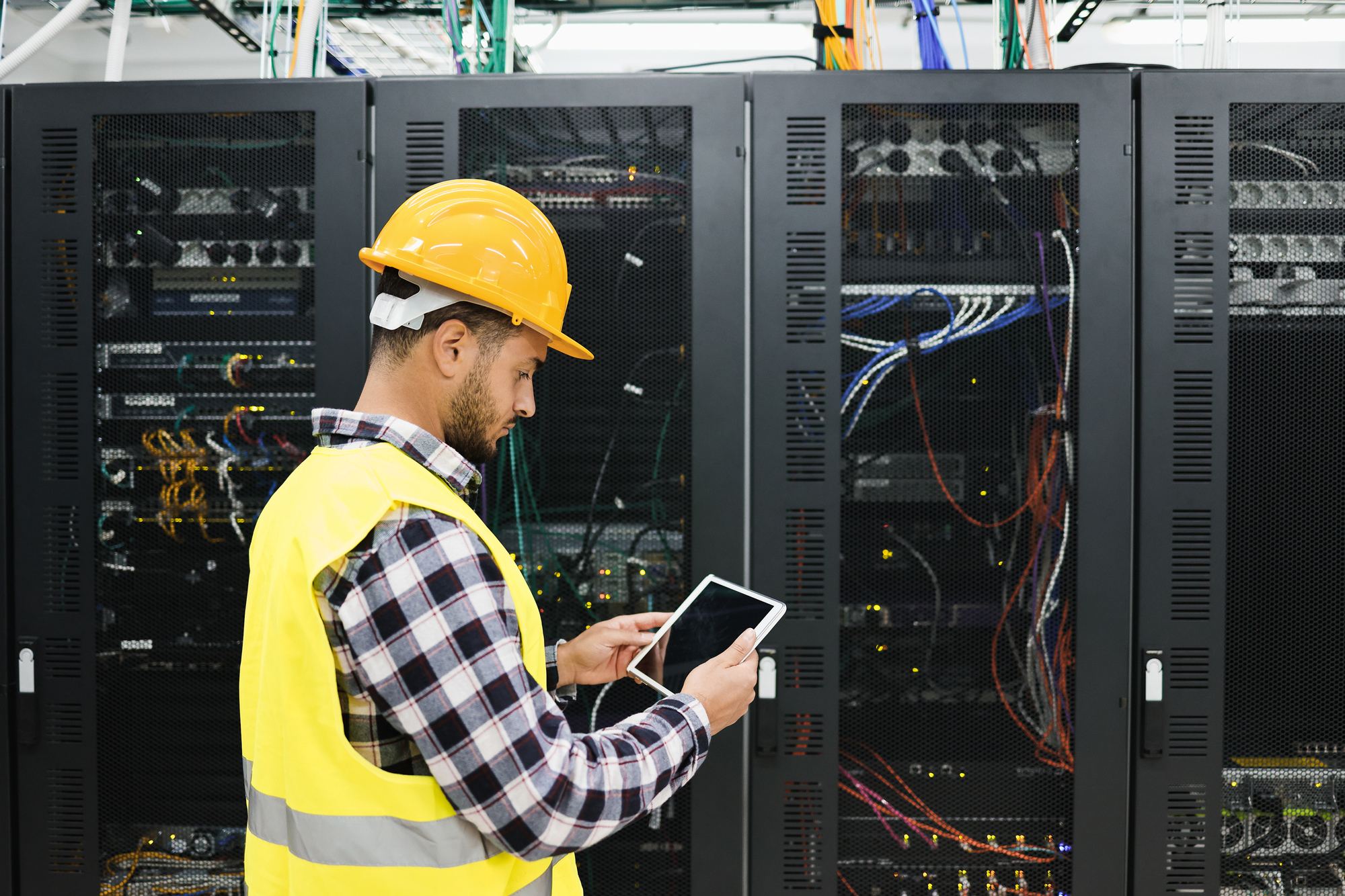 A worker in a hard hat and high-visibility vest checking a tablet beside server racks
